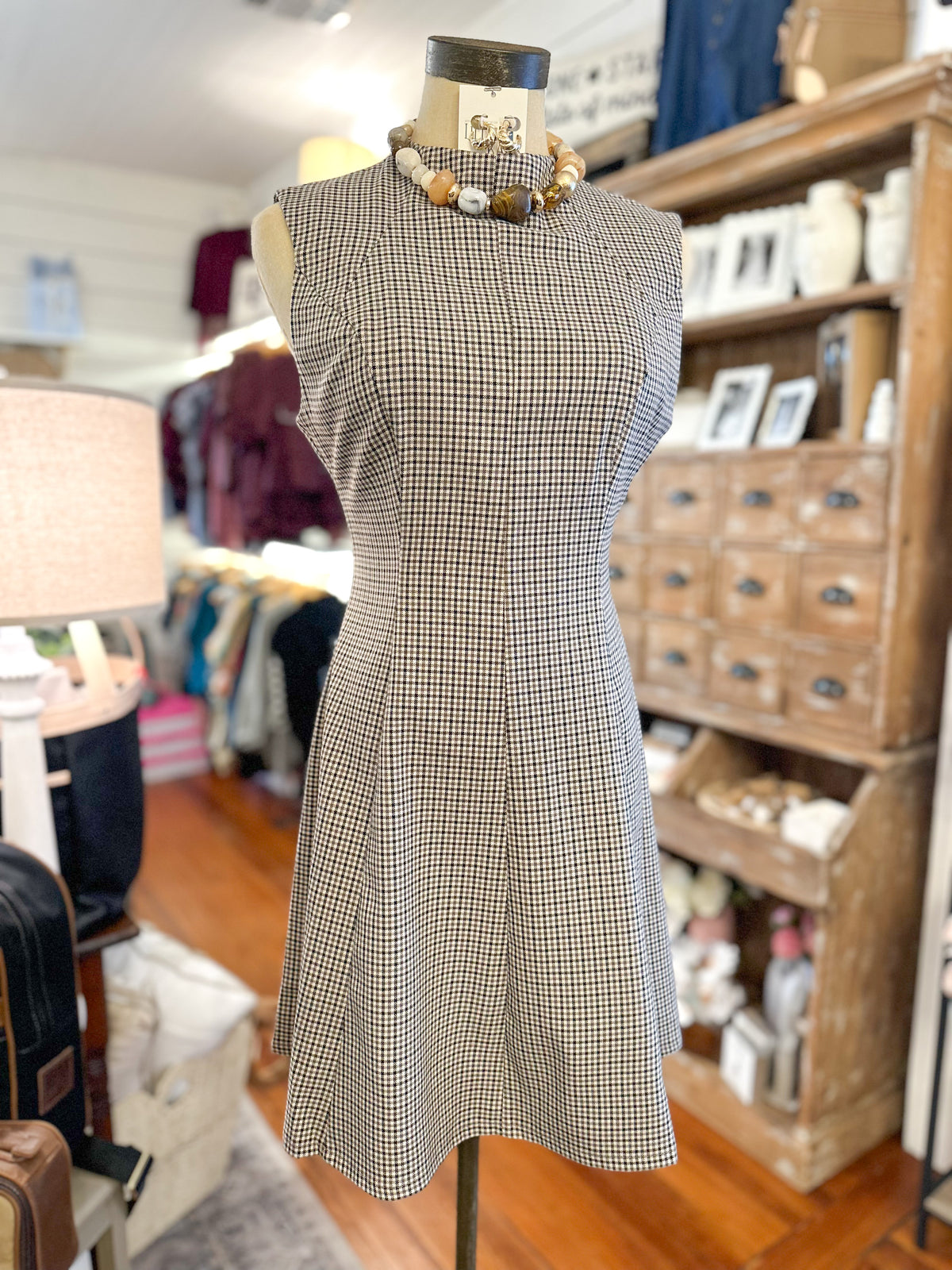 A sleeveless A-line dress with a small-scale black and beige check pattern, displayed on a mannequin torso in a retail shop setting.