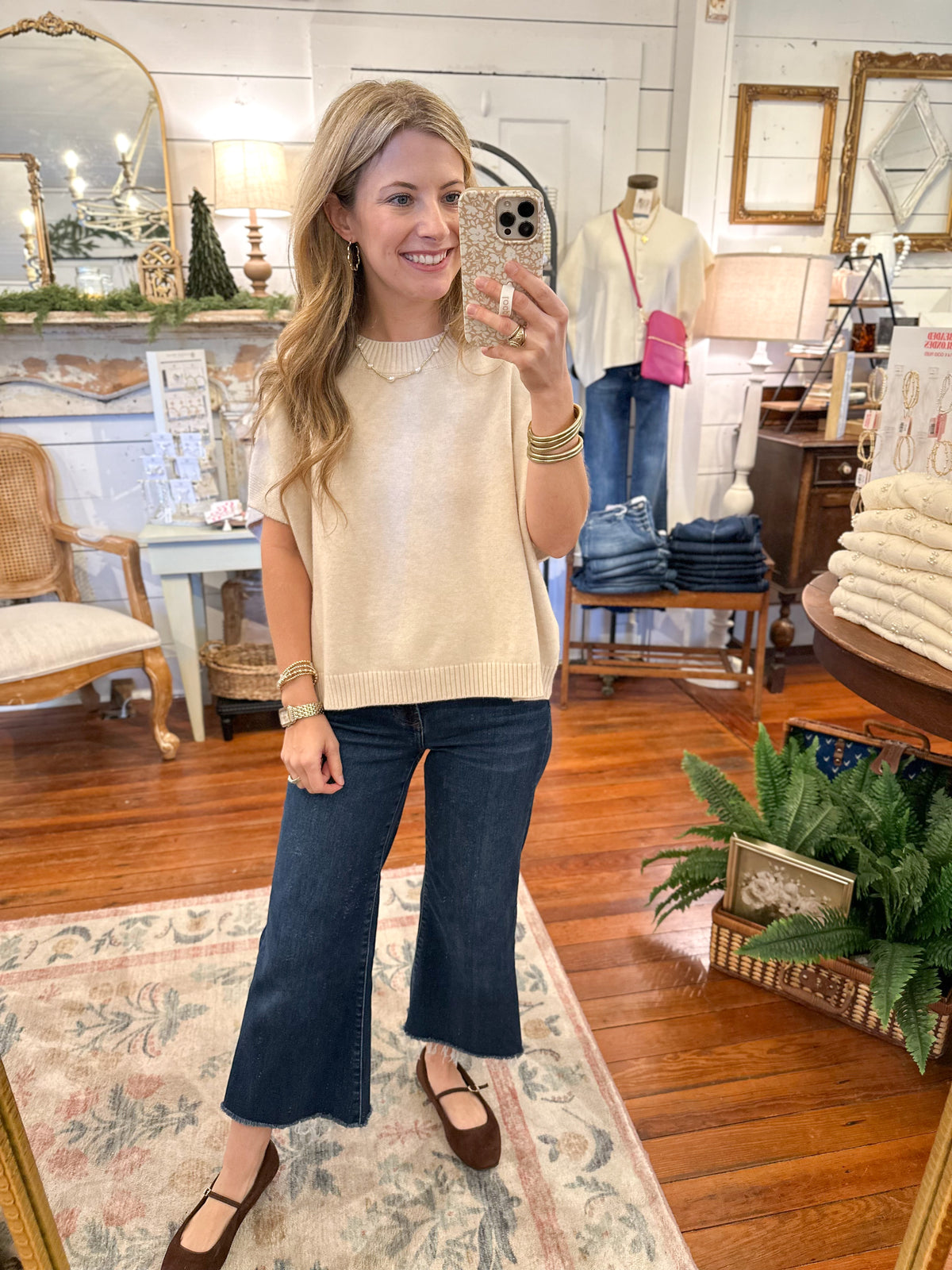 Woman taking a mirror selfie in a store, wearing a beige sweater and blue jeans.