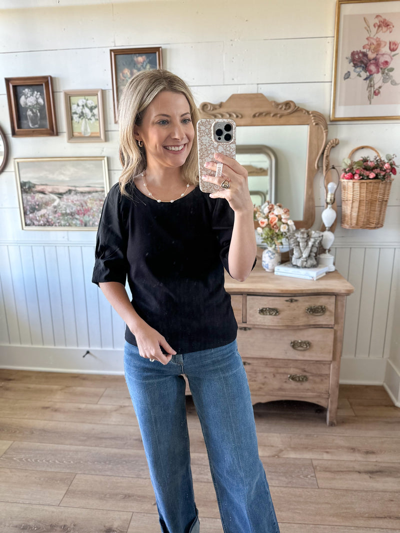 Woman taking a mirror selfie wearing a black top and blue jeans in a room with wooden furniture and decorative items.