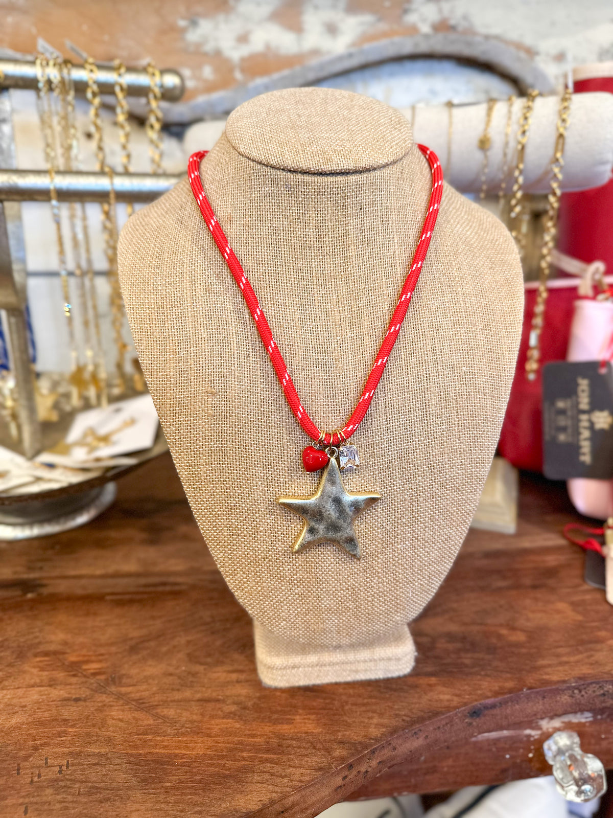 A red braided cord necklace with a silver-toned star pendant displayed on a beige fabric jewelry bust, set on a wooden surface with other jewelry in the background.