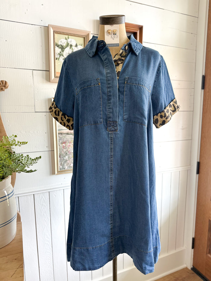 Blue denim dress with leopard print cuffs on a mannequin against a white paneled wall.