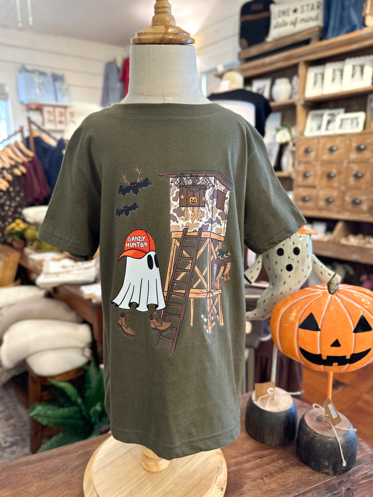 An olive green short-sleeve t-shirt displayed on a mannequin in a retail store. The back of the shirt features a graphic print of a haunted watchtower, a ghost, bats, and a pumpkin with the text 'Crazy Pockets'.