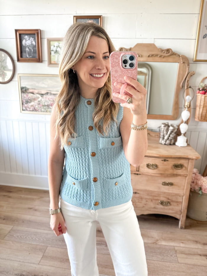 Woman taking a mirror selfie wearing a light blue knitted vest with brown buttons in a room.