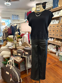 A mannequin in a clothing boutique wearing a black short-sleeve top with a lace hem and black wide-leg pants, styled with layered silver necklaces. The boutique interior features wood floors, shelves, and other clothing and accessories.