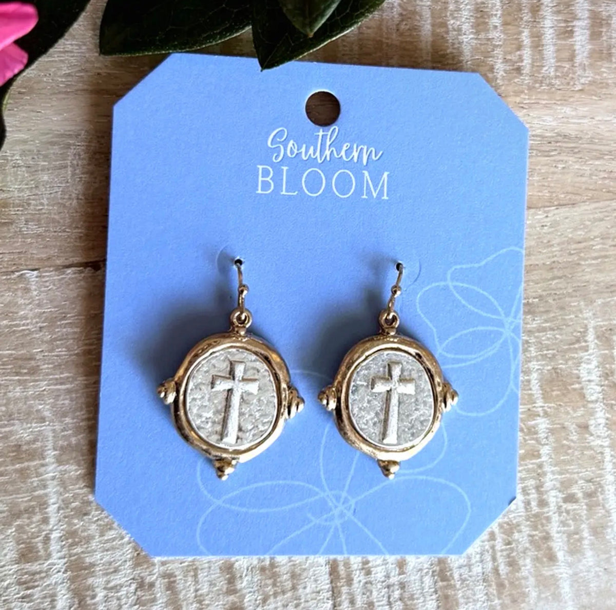 A pair of gold-toned round drop earrings with a raised cross motif on a textured light background, displayed on a blue card labeled Southern Bloom over a light wood surface.