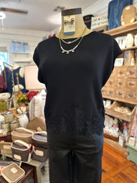 A black short-sleeve knit top with black lace trim at the hem, displayed on a mannequin torso in a boutique. The top is styled with layered gold necklaces 