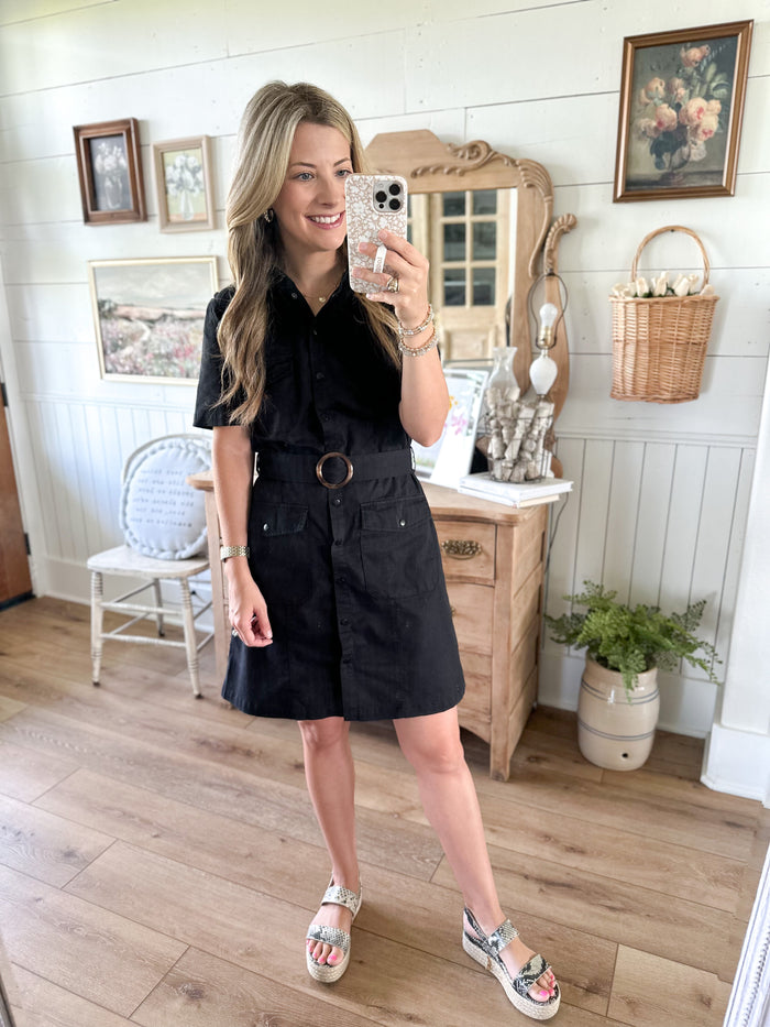 Woman taking a mirror selfie wearing a black dress in a room with wooden floor and white walls.