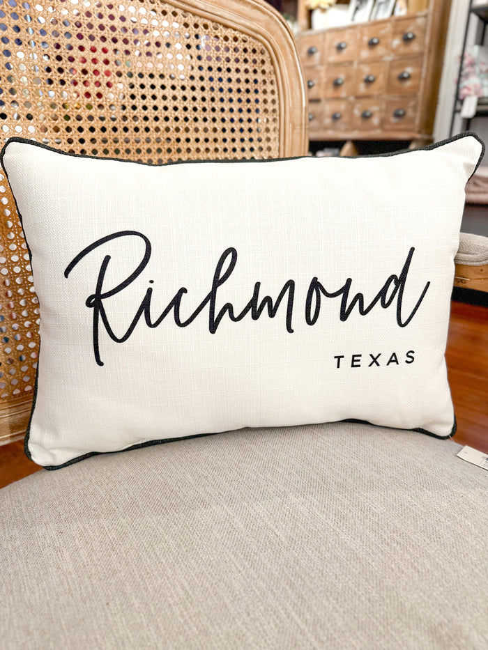 A rectangular cream throw pillow with black script reading 'Richmond' and smaller text 'TEXAS' underneath, resting on a beige upholstered chair in front of a woven cane chair.