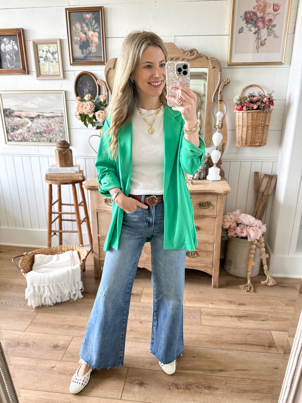 Woman taking a mirror selfie wearing a green cardigan, white top, and blue jeans in a home setting.