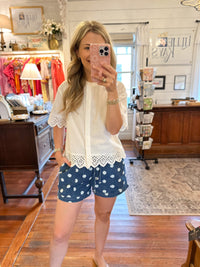 A woman taking a mirror selfie in a boutique shop, wearing a white short-sleeve blouse with lace trim and navy blue shorts with white polka dots.
