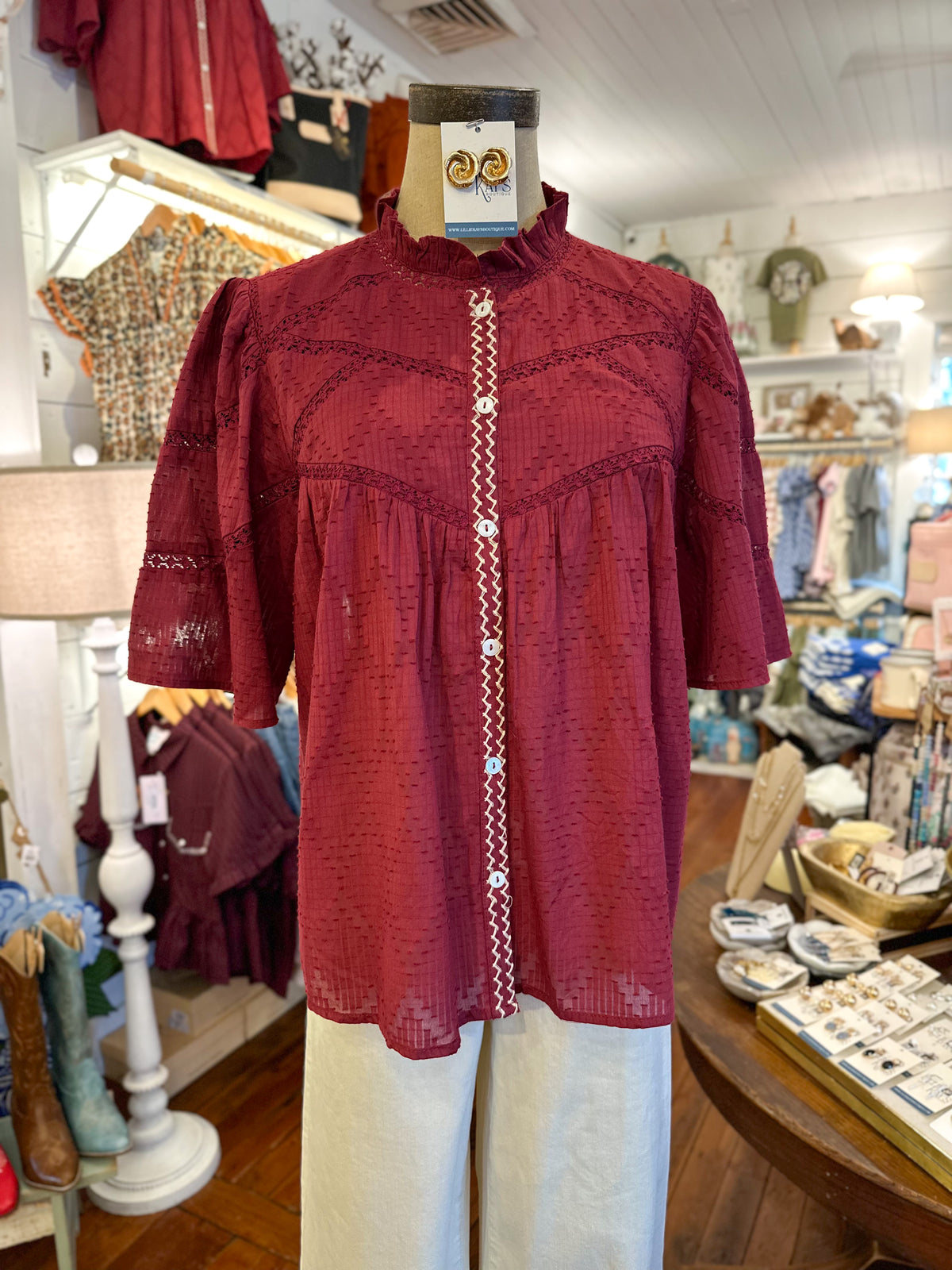 A burgundy short-sleeve blouse with a textured dot pattern and a decorative white embroidered vertical placket with white buttons, displayed on a mannequin in a retail store.