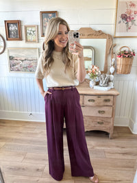 Woman taking a mirror selfie wearing a beige top and purple pants in a room with wooden floor and dresser.