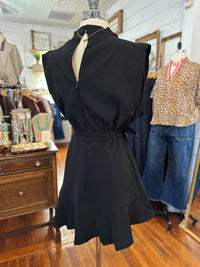 A sleeveless black dress displayed on a mannequin in a clothing boutique, featuring a high neckline with a zipper detail, a tied waist belt, and a flared ruffled hem.