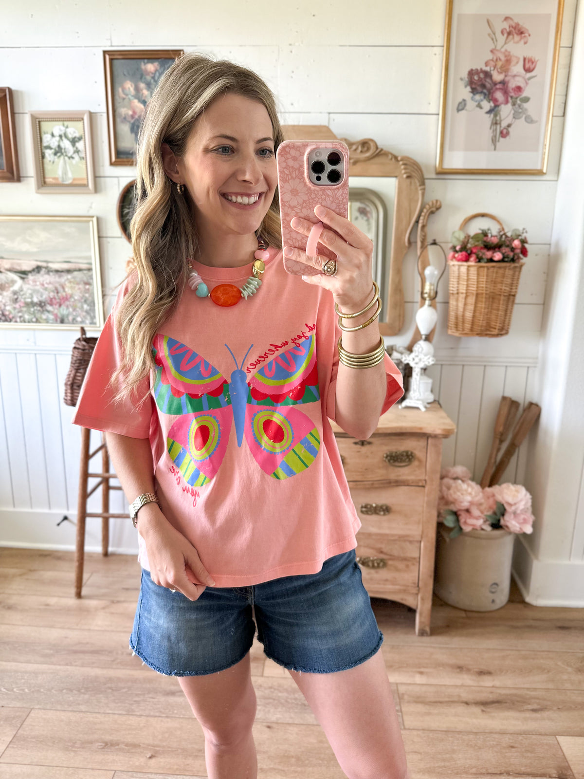 Woman taking a mirror selfie wearing a pink t-shirt with a colorful butterfly design and denim shorts in a home setting.