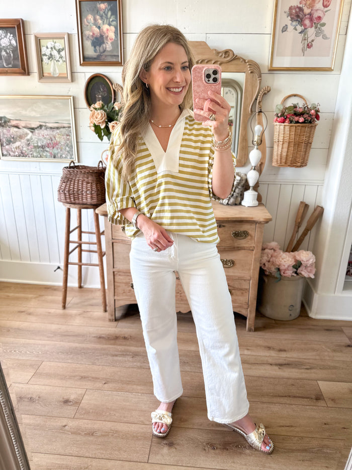 Woman taking a mirror selfie wearing a yellow and white striped shirt and white pants in a home setting.