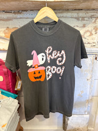 A dark gray graphic t-shirt hanging on a wooden hanger against a rustic distressed white wall. The shirt features a Halloween graphic with a jack-o'-lantern wearing a pink witch hat, a white ghost, and the text 'Hey BOO!'.
