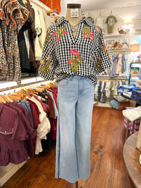 A black and white gingham blouse with pink floral embroidery and green cactus motifs, styled with light-wash blue wide-leg jeans on a mannequin inside a clothing boutique.