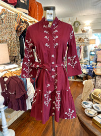 A burgundy long-sleeve button-front dress with white floral embroidery and a matching waist tie, displayed on a mannequin inside a clothing boutique.
