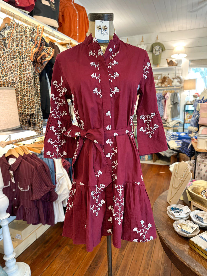 A burgundy long-sleeve button-front dress with white floral embroidery and a matching waist tie, displayed on a mannequin inside a clothing boutique.