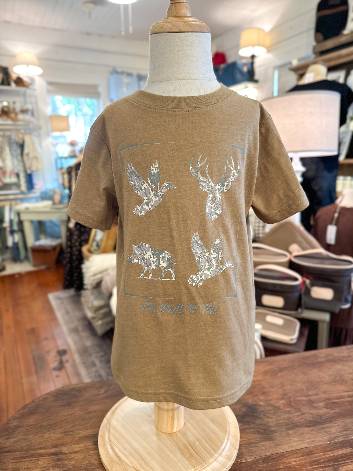 A toddler's short-sleeve t-shirt in a warm tan color, displayed on a wooden mannequin torso inside a retail store.