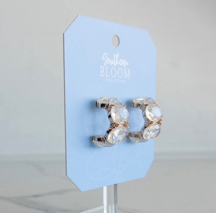 A pair of gold crescent-shaped hoop earrings with clear faceted gemstones, displayed on a light blue card from Southern Bloom Collections.