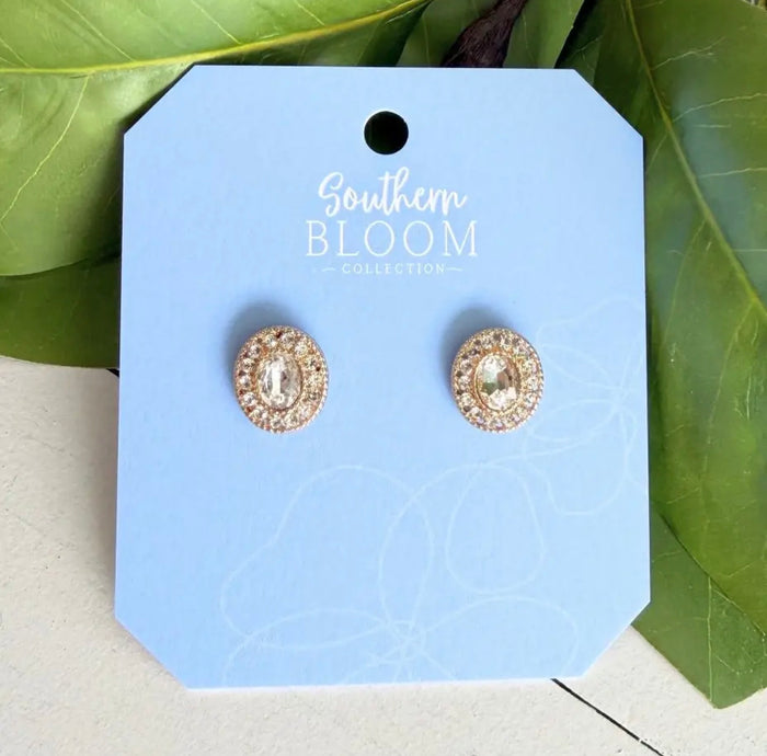 A pair of round stud earrings with gold-toned crystal clusters, displayed on a light blue card labeled Southern Bloom Collection, set against a background of green leaves.
