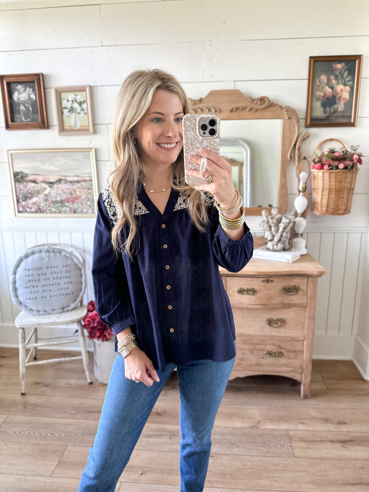 Woman taking a mirror selfie wearing a navy blue shirt and blue jeans in a room with wooden furniture and decorative items.