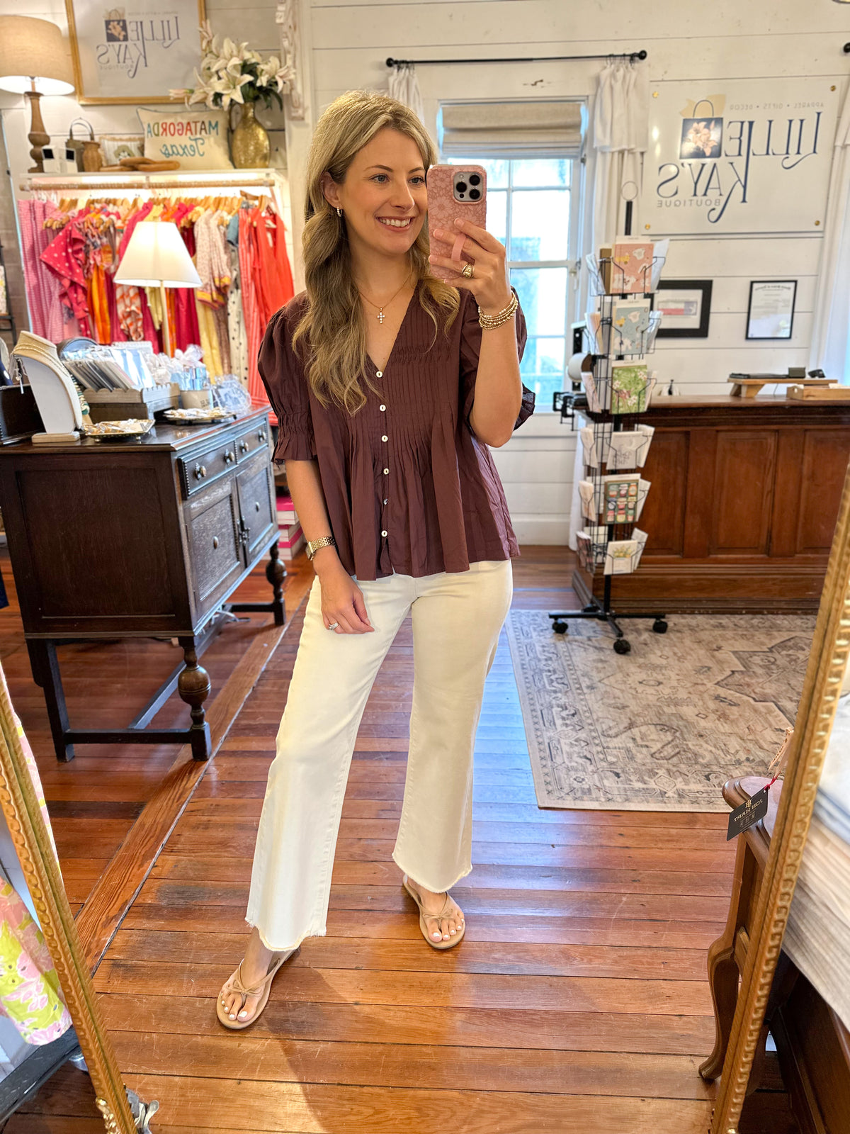 A woman takes a mirror selfie in a boutique while wearing a short-sleeve button-up blouse in a dusty mauve-brown color with white wide-leg pants and beige flat sandals.