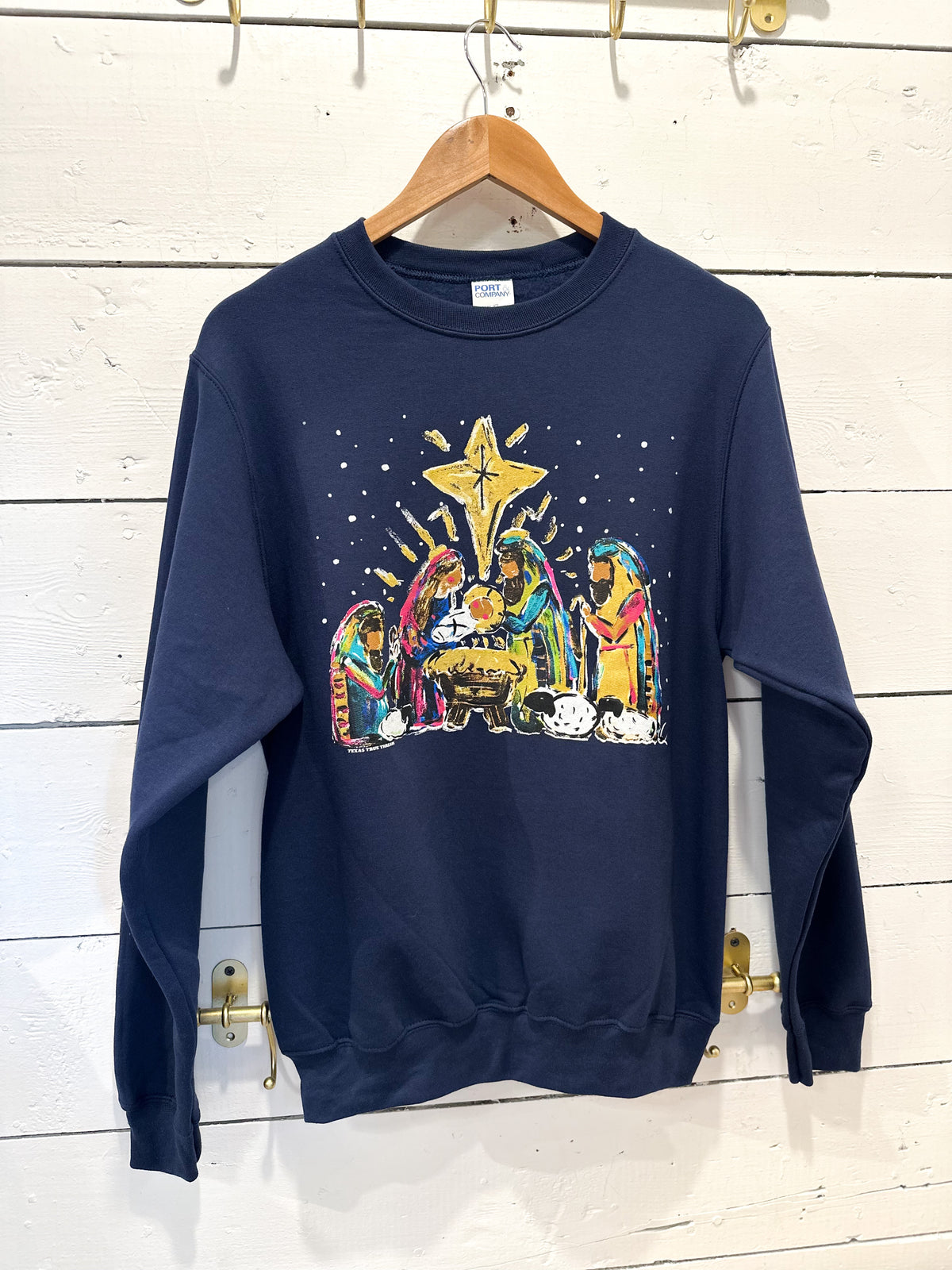 Navy blue sweatshirt with a colorful nativity scene design on a wooden hanger against a white wooden panel background.