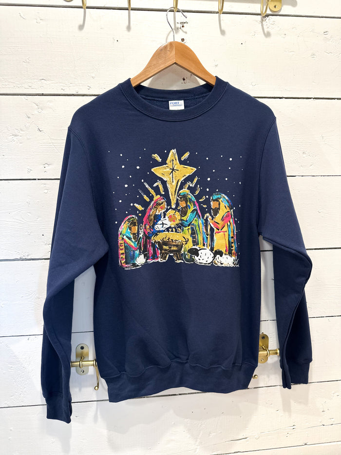 Navy blue sweatshirt with a colorful nativity scene design on a wooden hanger against a white wooden panel background.
