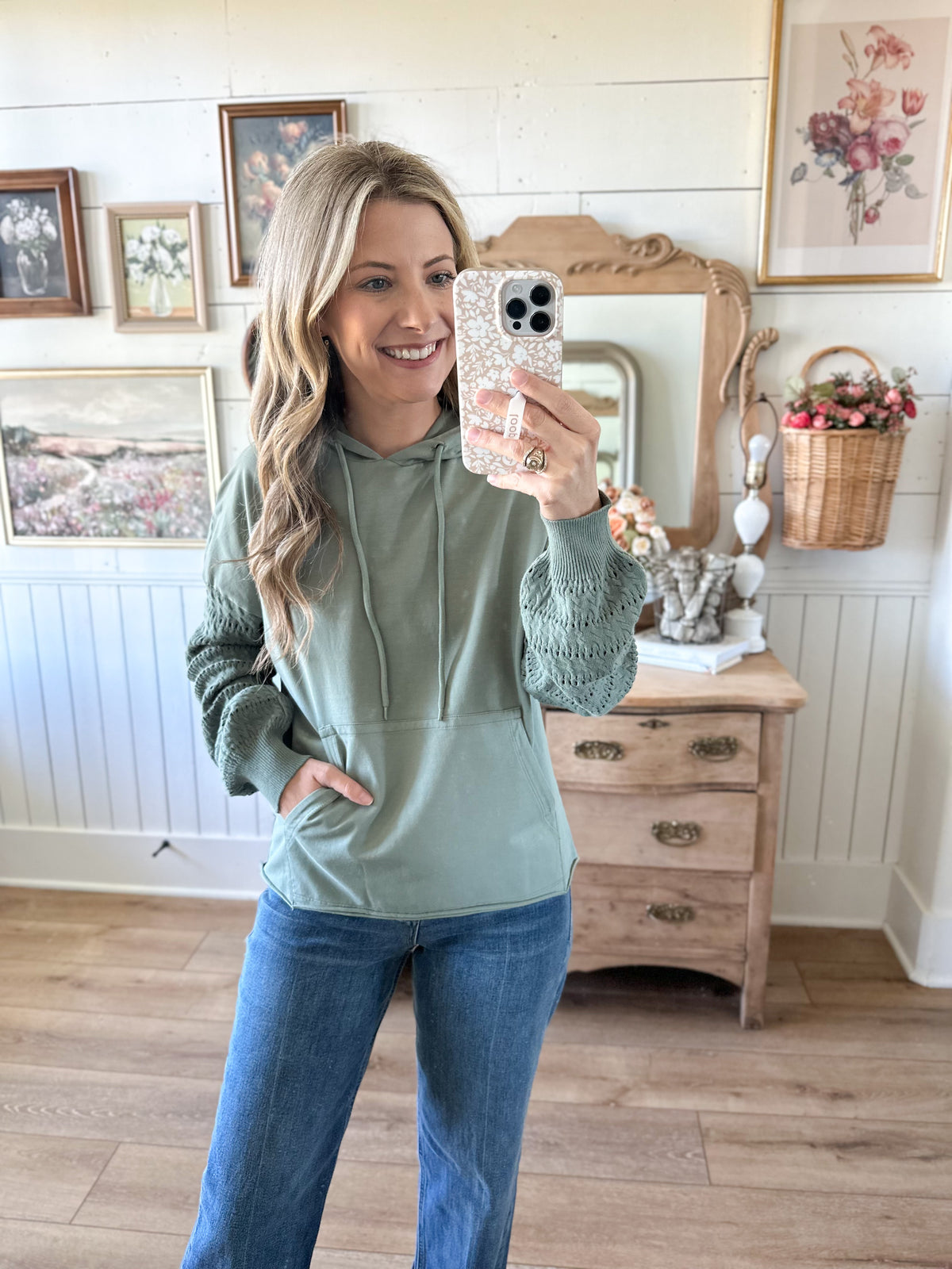 Woman taking a mirror selfie wearing a green hoodie and blue jeans in a room with wooden flooring and framed pictures on the wall.