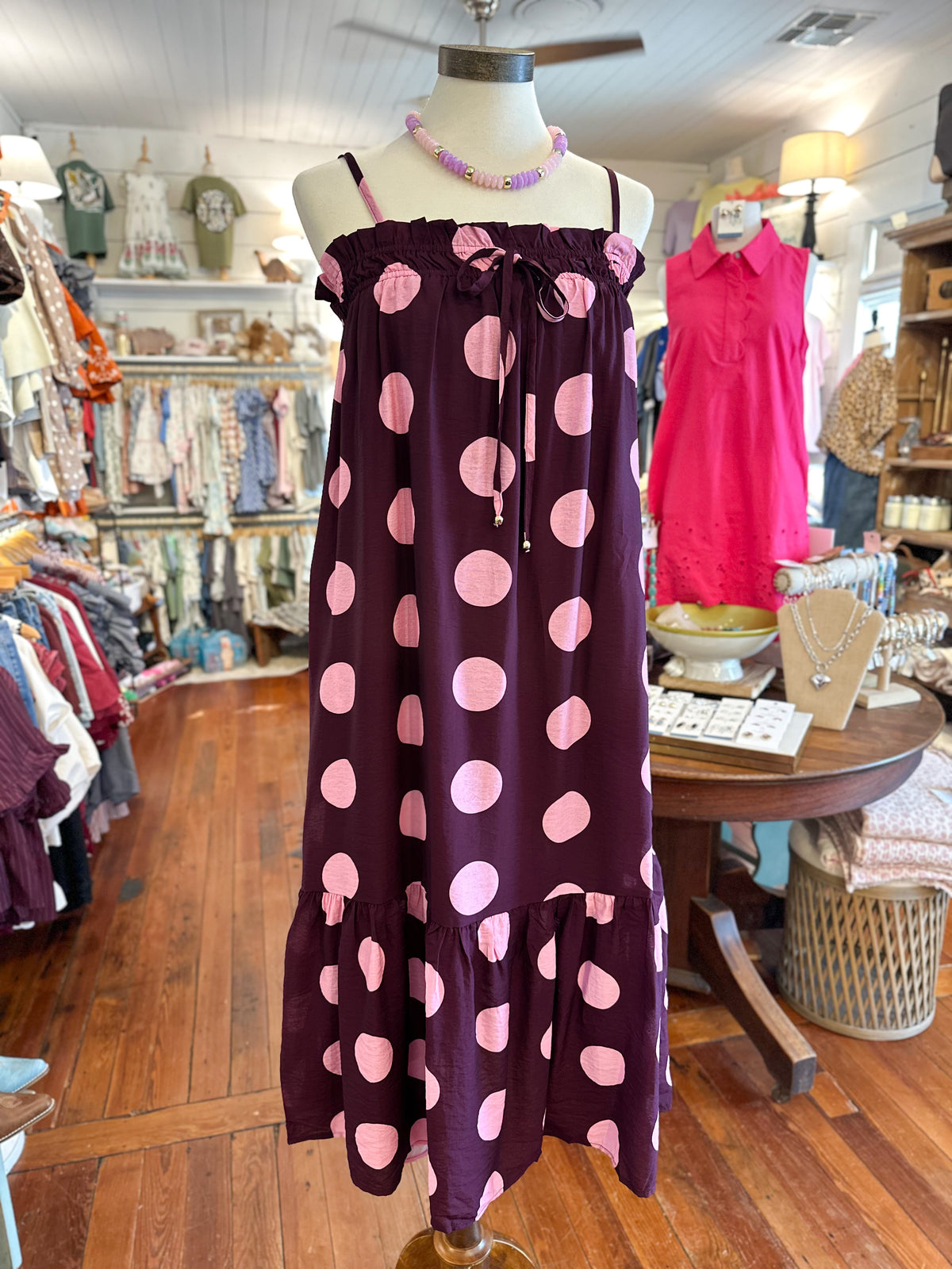 A sleeveless tiered maxi dress with a dark purple base and large light pink polka dots, displayed on a mannequin inside a clothing boutique.