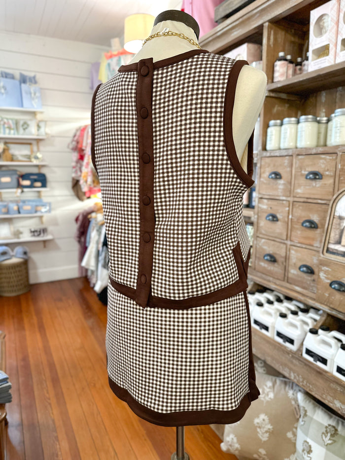 A sleeveless brown and beige gingham top displayed on a mannequin in a retail shop. The back view shows a brown button placket and matching brown trim.