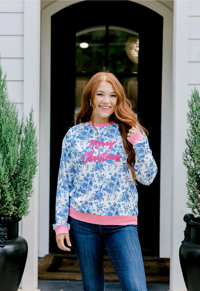 Woman wearing a floral sweatshirt with 'Merry Christmas' text, standing in front of a door.