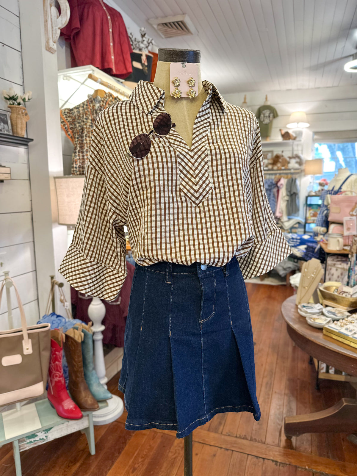 A mannequin in a boutique displays a long-sleeve brown and beige gingham button-up shirt paired with a dark blue denim skirt.