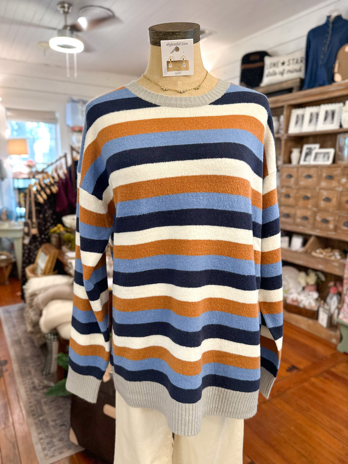 A striped crewneck sweater displayed on a mannequin torso inside a retail store. The sweater features horizontal stripes in navy blue, light blue, rust orange, and cream, with a solid gray ribbed crew neck, cuffs, and hem.