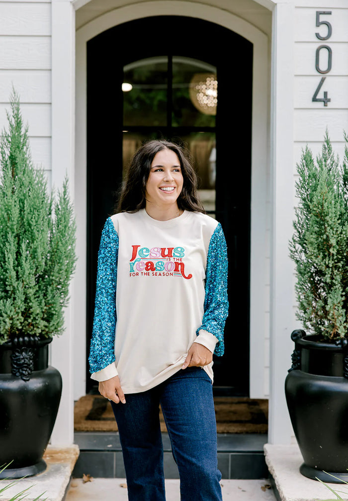 Woman wearing a festive shirt with Jesus is the reason for the season standing in front of a house entrance.