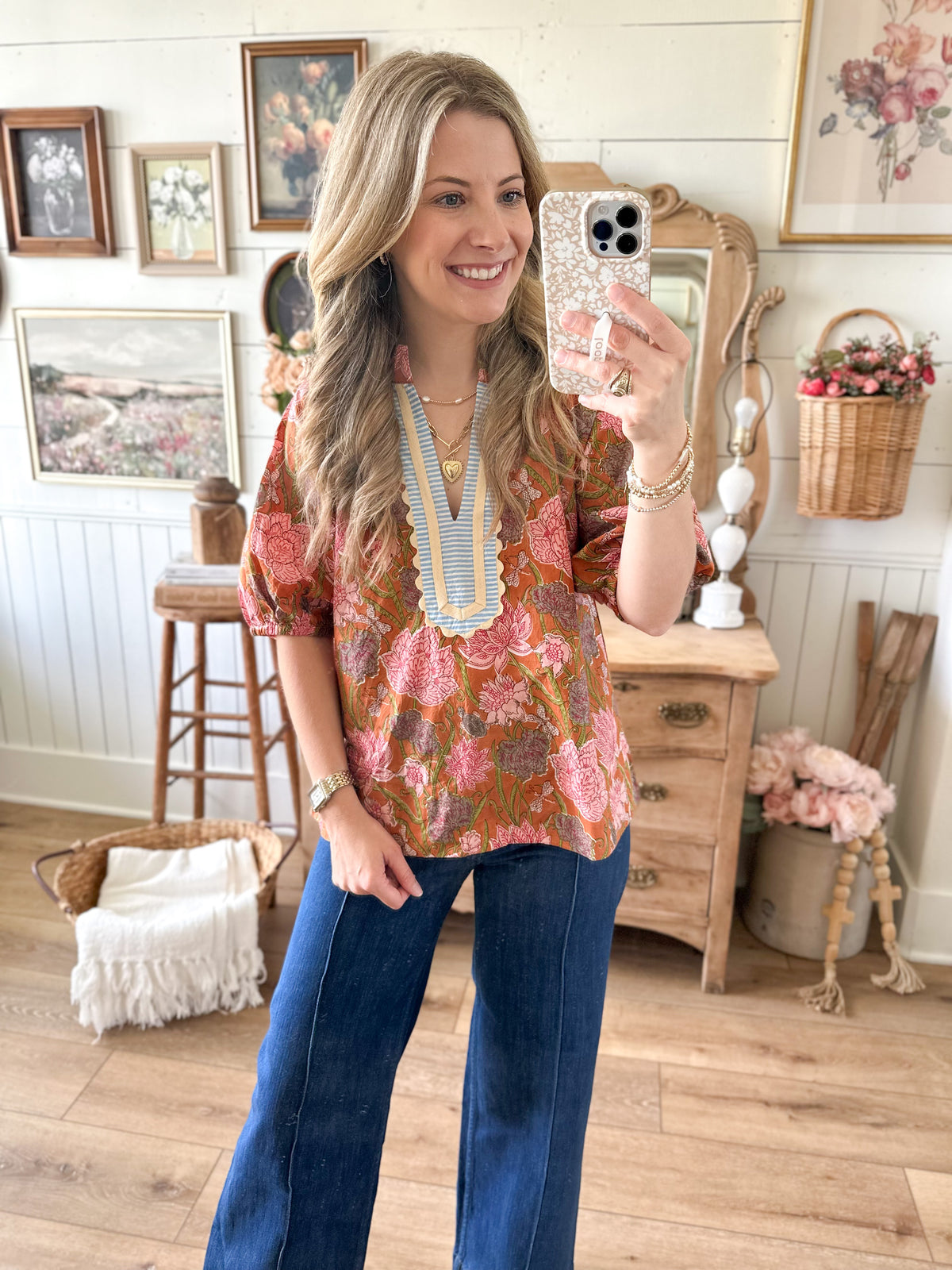 Woman taking a mirror selfie wearing a floral top and blue jeans in a home setting.