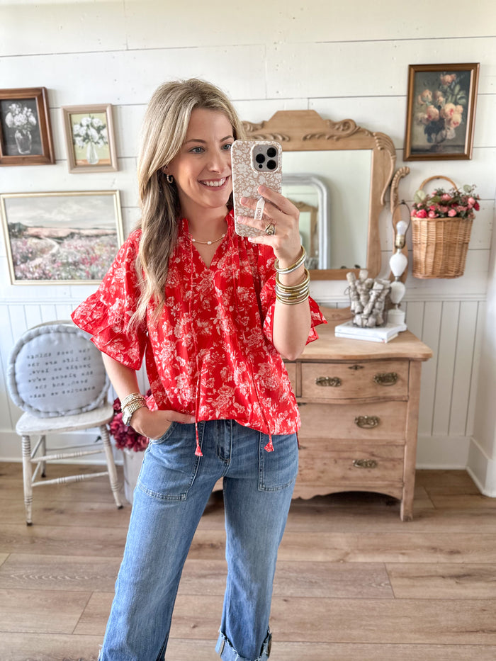 Woman taking a mirror selfie wearing a red floral blouse and blue jeans in a room with wooden furniture and framed pictures.