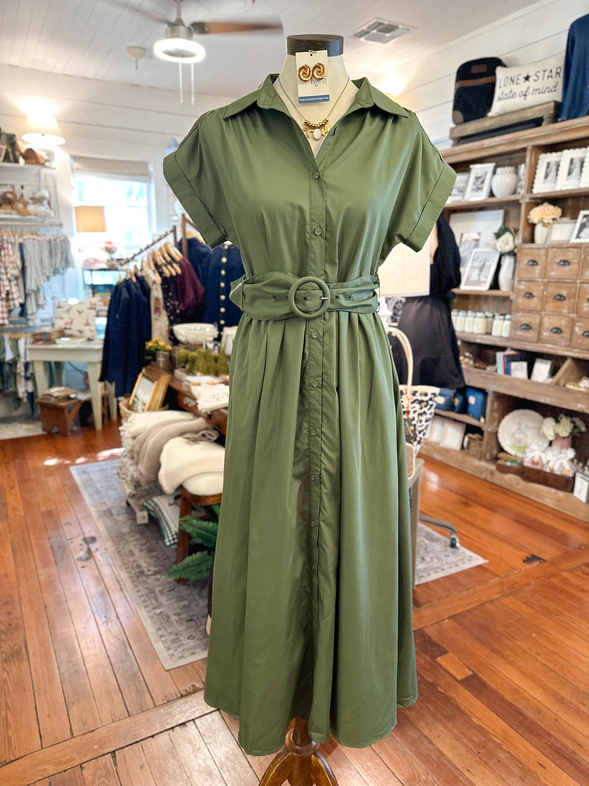 An olive green short-sleeve shirt dress displayed on a mannequin in a boutique. The dress features a collared neckline, button-front bodice, a matching fabric belt with a large round buckle, and a gathered skirt.