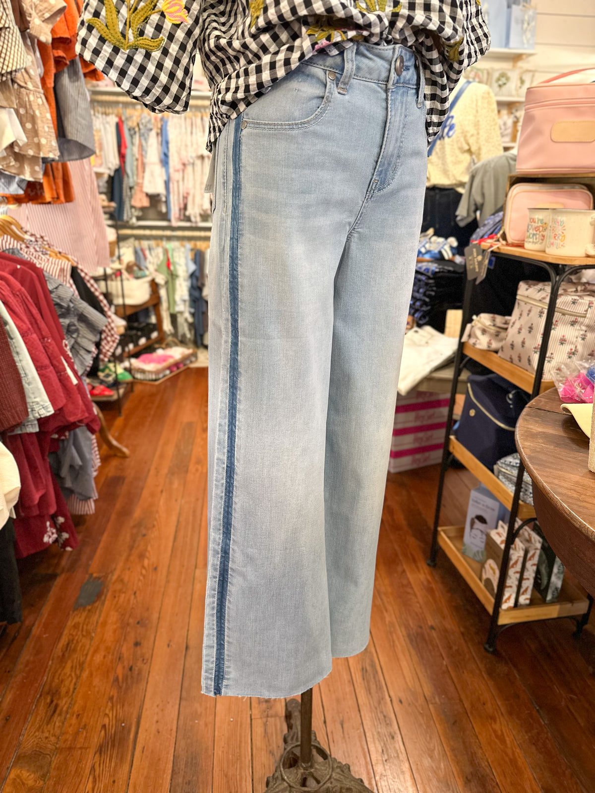 Light wash wide-leg denim jeans displayed on a mannequin in a clothing boutique, featuring a blue stripe down the side of the leg.