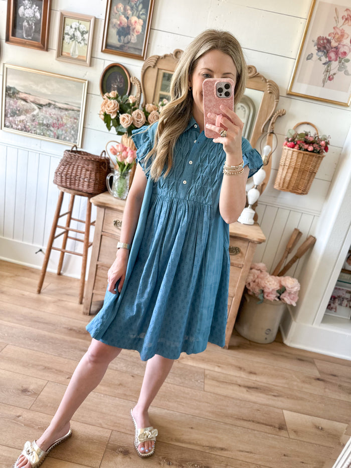 Woman taking a mirror selfie wearing a blue dress in a home setting with wooden floors and decorative items.