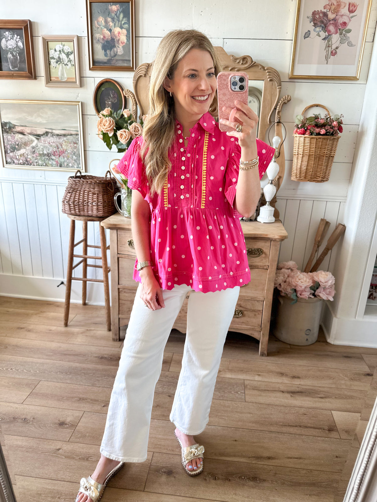 Woman taking a mirror selfie wearing a pink blouse and white pants in a home setting.