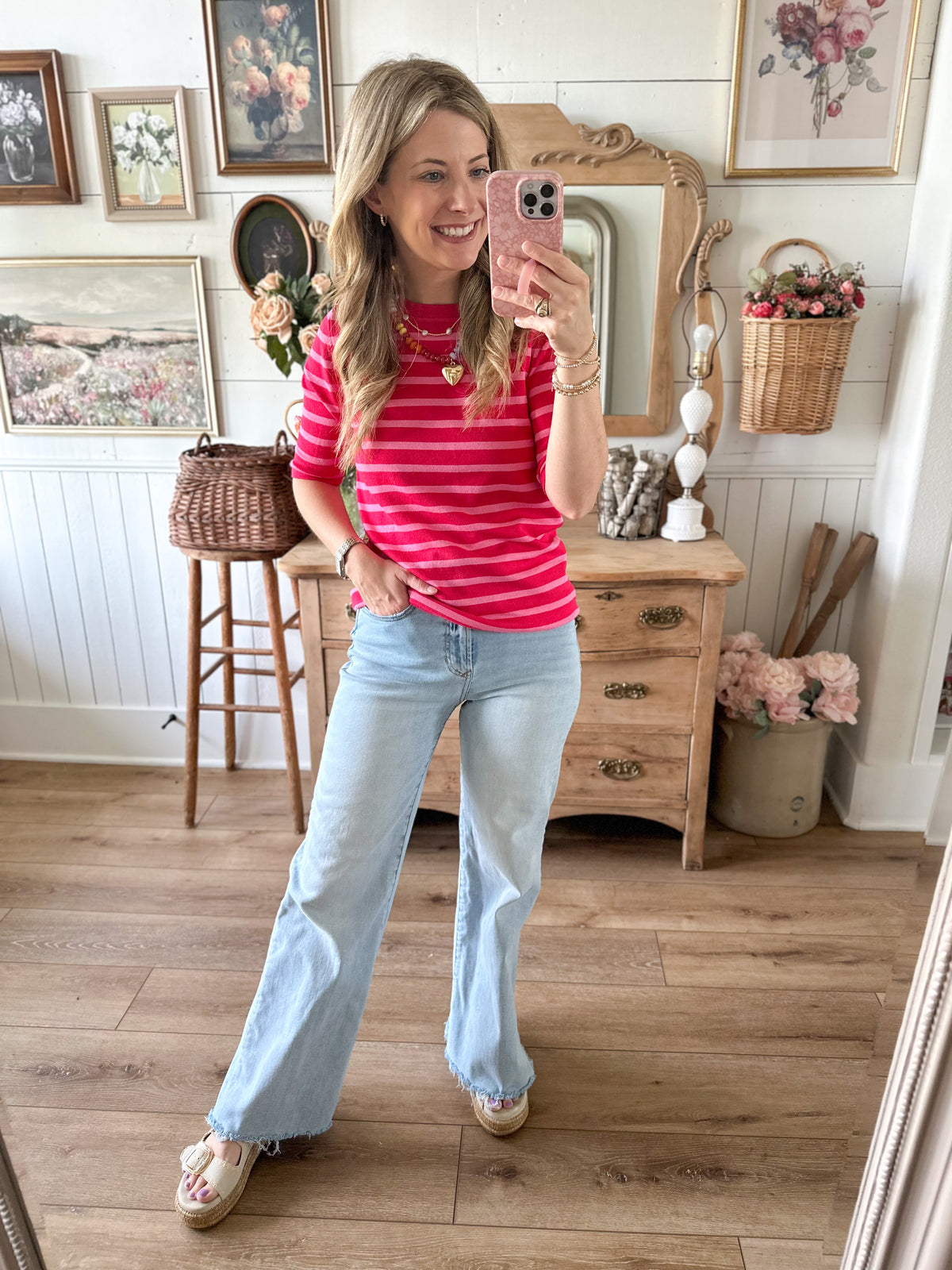 Woman taking a mirror selfie wearing a pink striped shirt and light blue jeans in a home setting.