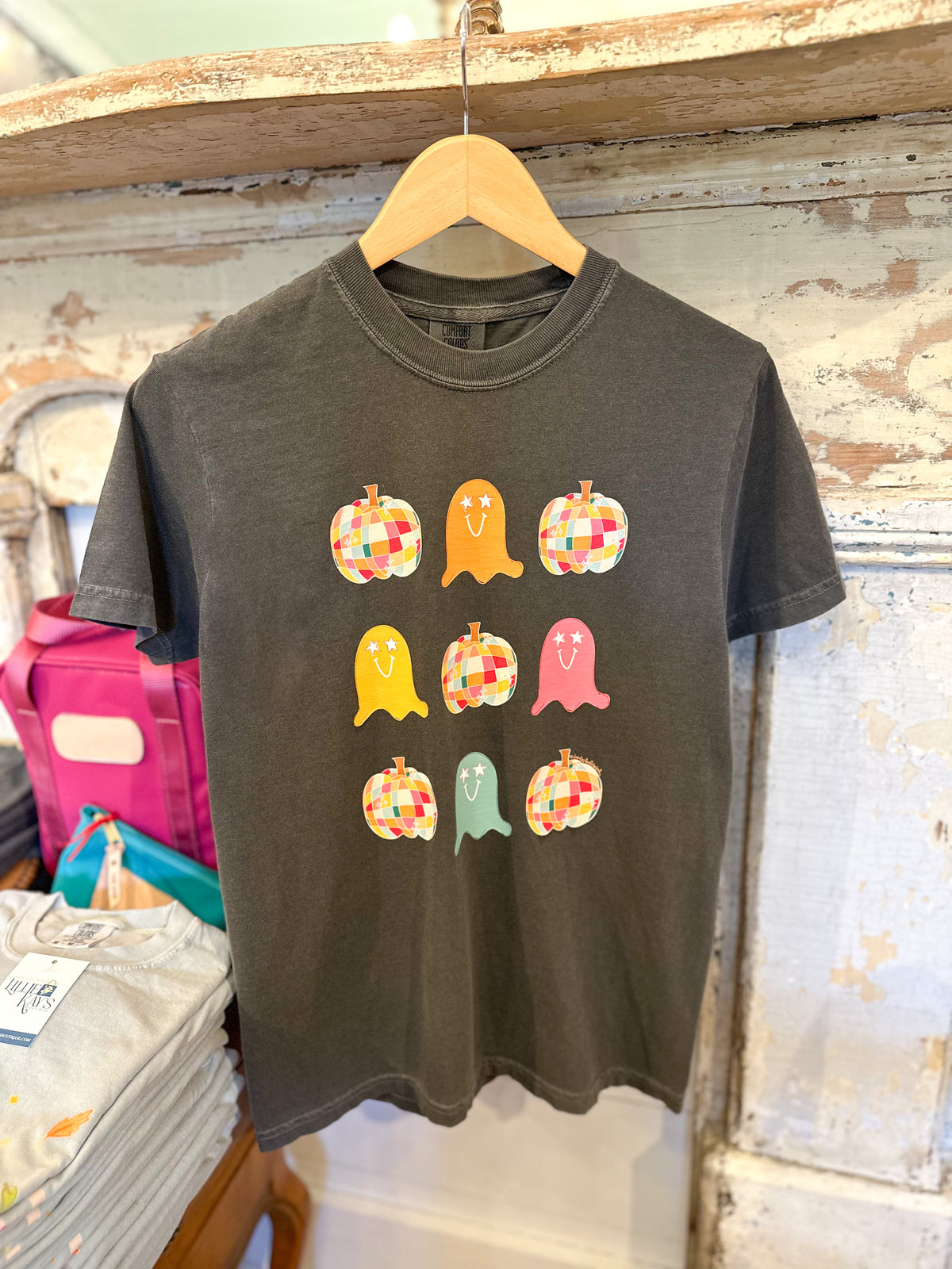 A dark charcoal gray t-shirt hanging on a wooden hanger against a rustic distressed wood wall. The shirt features a graphic of nine colorful patchwork-style pumpkins and ghosts in orange, yellow, pink, green, and brown.