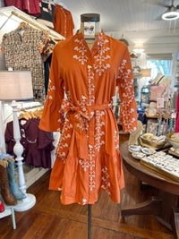 An orange-brown short dress with white floral embroidery and a matching tie belt, displayed on a mannequin inside a clothing boutique.