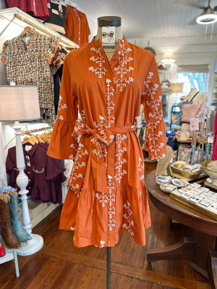 An orange-brown short dress with white floral embroidery and a matching tie belt, displayed on a mannequin inside a clothing boutique.
