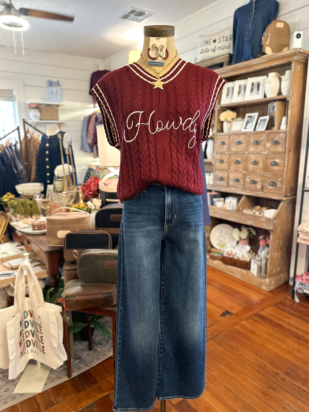A mannequin in a rustic clothing boutique displays a burgundy cable-knit sweater vest with 'Howdy!' embroidered in white script on the chest, paired with jeans.