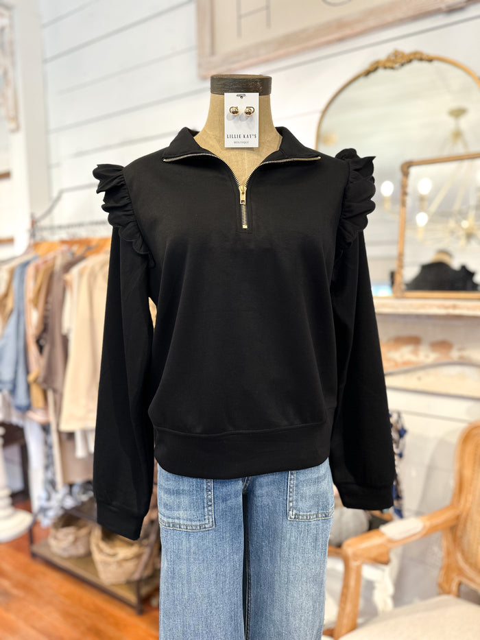 Black pullover with ruffled shoulders on a mannequin in a store setting.