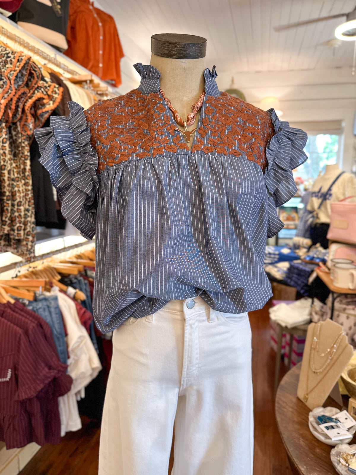 A blue and white vertically striped blouse with a rust-orange patterned embroidered detail on chest and ruffled sleeves, displayed on a mannequin torso in a clothing boutique.